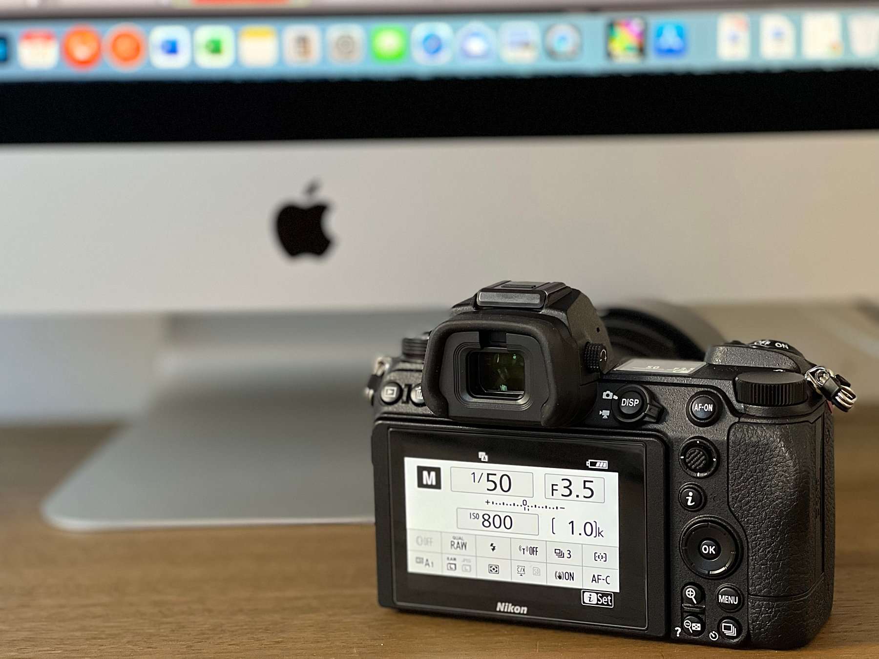 A Nikon camera displaying manual settings including shutter speed, aperture, and ISO, placed in front of an Apple iMac. This is illustrating how understanding the exposure triangle for portrait photographers can help capture balanced images.