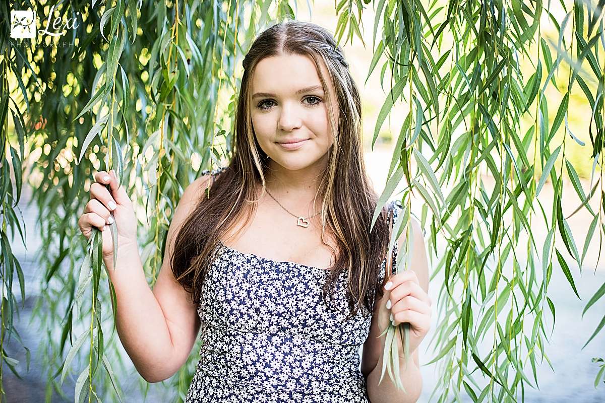 A young woman stands gently holding hanging willow tree branches, using the draped foliage for soft natural framing.