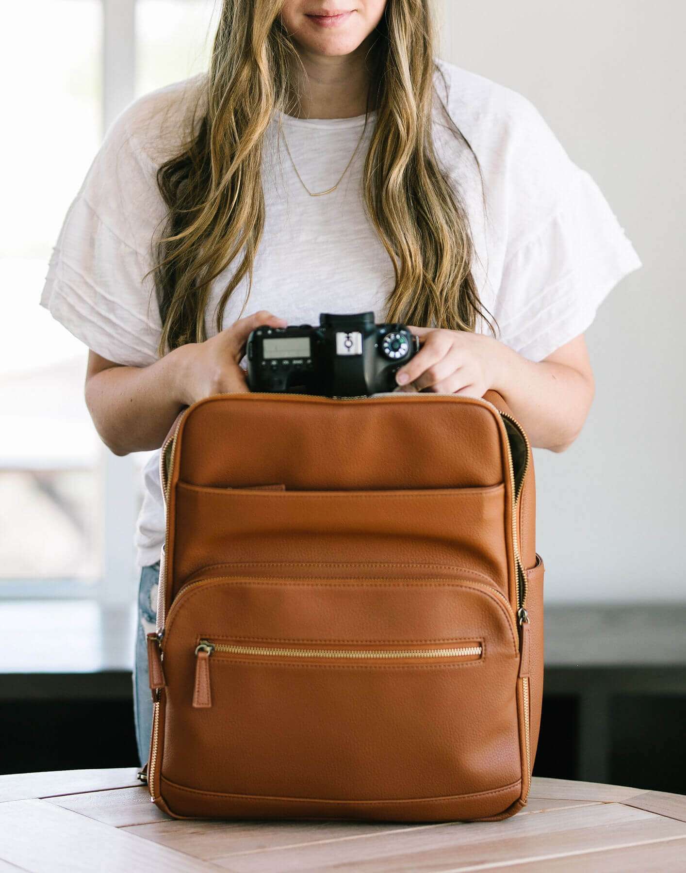 A woman in a white tee is placing a camera into a stylish tan leather backpack.