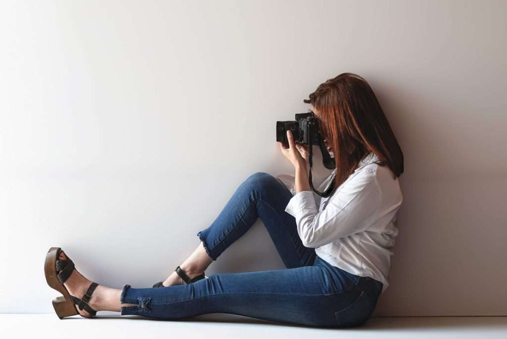 Female photographer sitting on the floor and holding a camera up to her eye, demonstrating a stable shooting position to avoid blur created by using a lower shutter speed.