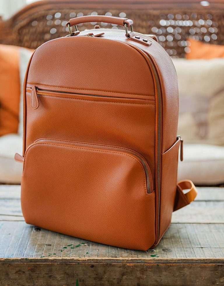 Brown leather backpack style camera bag sitting on a rustic wooden table. The structured bag has multiple zippered pockets and a top handle designed to organize gear, making it a practical option among stylish camera bags for women.