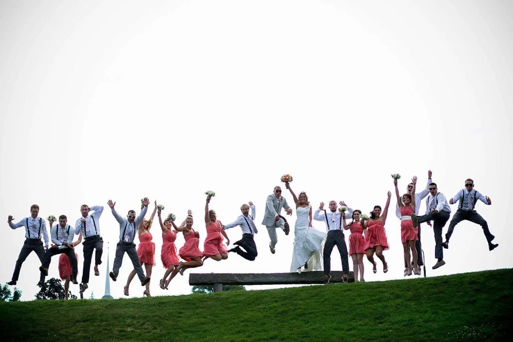 Group of wedding party members in formal attire jumping in the air on a grassy hill with arms raised against a bright sky, demonstrating fast shutter speed freezing motion for camera settings for beginners.