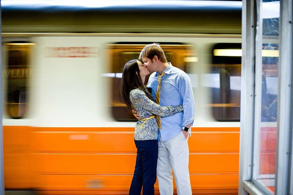 Couple kissing on a train platform while an orange train blurs behind them in motion, illustrating slow shutter speed creating motion blur in camera settings for beginners.